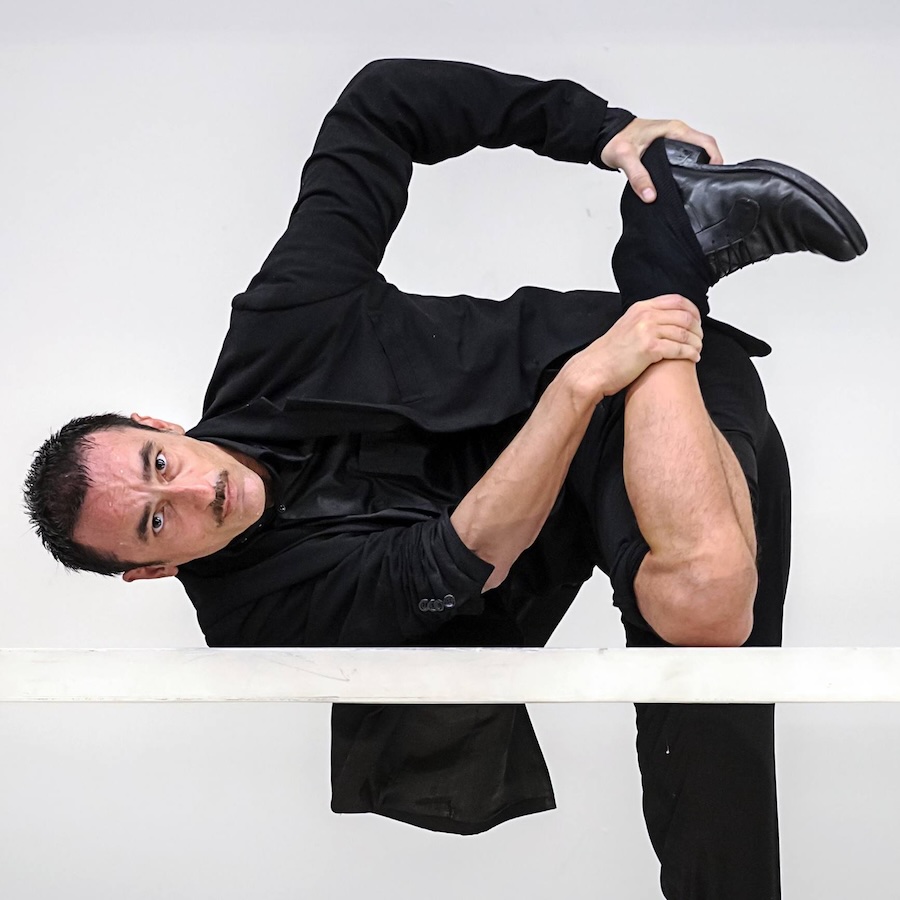 Artist Dressed in black white background  posed with one leg down and holding one foot up